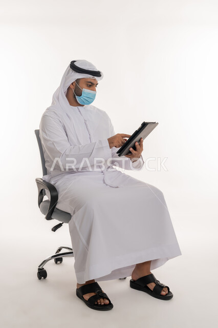 مفهوم العمل عن بعد للحفاظ على السلامة, القيام بالإجراءات الاحترازية للوقاية من كوفيد 19, استخدام الأجهزة التقنية الحديثة لنشر الوعي الصحي والحد من انتشار الأمراض, بورتريه لرجل عربي خليجي اماراتي يرتدي الزي التقليدي يضع الكمامة يعمل على الجهاز اللوحي, خلفية بيضاء
