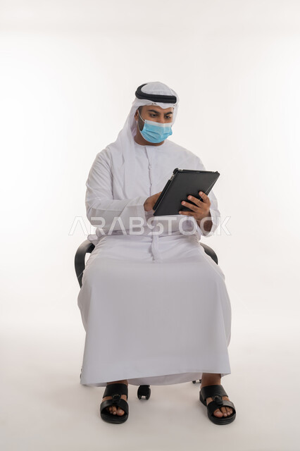 القيام بالإجراءات الاحترازية للوقاية من كوفيد 19, استخدام الأجهزة التقنية الحديثة لنشر الوعي الصحي والحد من انتشار الأمراض, مفهوم العمل عن بعد للحفاظ على السلامة, بورتريه لرجل عربي خليجي اماراتي يرتدي الزي التقليدي يضع الكمامة يعمل على الجهاز اللوحي, خلفية بيضاء