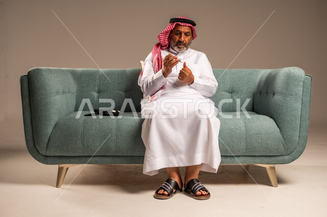جهاز إلكتروني حديث لقياس سكر الدم لمرضى داء السكري، بورتريه لرجل مسن  عربي خليجي سعودي مريض يرتدي الشماغ والثوب التقليدي يجلس على كنبة مريحة في غرفة المعيشة يقوم بفحص نسبة السكر في دمه، الاهتمام والرعاية بالصحة الجسدية، خلفية بيج