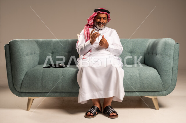 جهاز إلكتروني حديث لقياس سكر الدم لمرضى داء السكري، بورتريه لرجل مسن  عربي خليجي سعودي مريض يرتدي الشماغ والثوب التقليدي يجلس على كنبة مريحة في غرفة المعيشة يقوم بفحص نسبة السكر في دمه، الاهتمام والرعاية بالصحة الجسدية، خلفية بيج