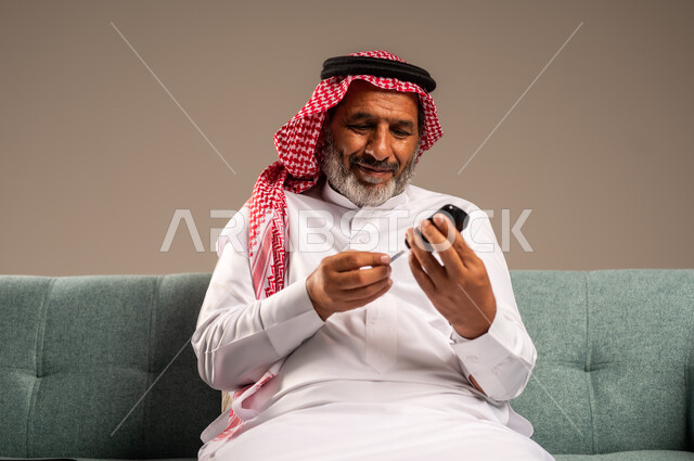 جهاز إلكتروني حديث لقياس سكر الدم لمرضى داء السكري، بورتريه لرجل مسن  عربي خليجي سعودي مريض يرتدي الشماغ والثوب التقليدي يجلس على كنبة مريحة في غرفة المعيشة يقوم بفحص نسبة السكر في دمه، الاهتمام والرعاية بالصحة الجسدية، خلفية بيج