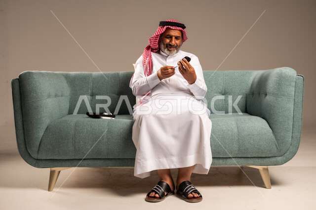 جهاز إلكتروني حديث لقياس سكر الدم لمرضى داء السكري، بورتريه لرجل مسن  عربي خليجي سعودي مريض يرتدي الشماغ والثوب التقليدي يجلس على كنبة مريحة في غرفة المعيشة يقوم بفحص نسبة السكر في دمه، الاهتمام والرعاية بالصحة الجسدية، خلفية بيج