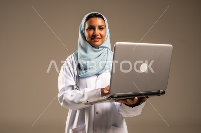 استخدام التكنولوجيا في البحث والتطوير، تقديم الخدمات للمرضى بكل كفاءه، اجراء الاستشارات الطبيه عن بعد، بورتريه لطبيبة عربية خليجية سعودية ترتدي عباءة ملونة مع المعطف الابيض تعمل على جهاز الحاسوب المحمول، مهن ووظائف صحية، خلفيه ملونة
