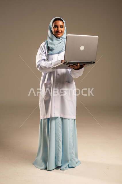 استخدام التكنولوجيا في البحث والتطوير، تقديم الخدمات للمرضى بكل كفاءه، اجراء الاستشارات الطبيه عن بعد، بورتريه لطبيبة عربية خليجية سعودية ترتدي عباءة ملونة مع المعطف الابيض تعمل على جهاز الحاسوب المحمول، مهن ووظائف صحية، خلفيه ملونة