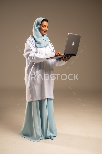 استخدام التكنولوجيا في البحث والتطوير، تقديم الخدمات للمرضى بكل كفاءه، اجراء الاستشارات الطبيه عن بعد، بورتريه لطبيبة عربية خليجية سعودية ترتدي عباءة ملونة مع المعطف الابيض تعمل على جهاز الحاسوب المحمول، مهن ووظائف صحية، خلفيه ملونة