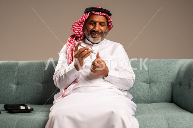الرعاية الصحية والفحص الطبي السريع في المنزل، جهاز إلكتروني حديث لمرضى داء السكري، بورتريه لرجل كبير بالسن عربي خليجي سعودي مريض يرتدي الشماغ والثوب التقليدي يجلي على كنبة مريحة يقوم بفحص نسبة السكر في دمه، ،خلفية بيج