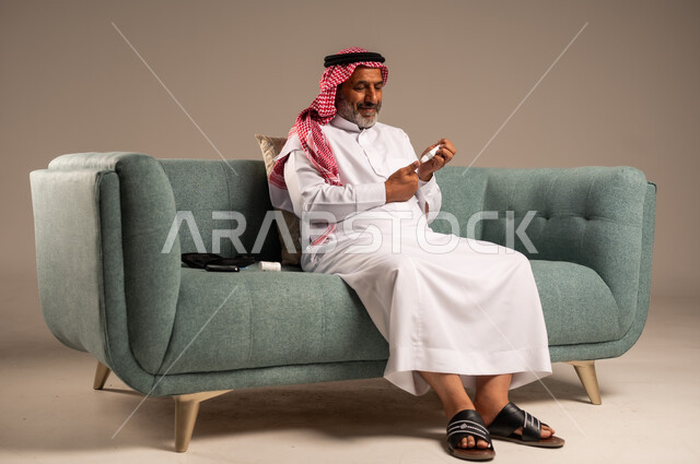 الرعاية الصحية والفحص الطبي السريع في المنزل، جهاز إلكتروني حديث لمرضى داء السكري، بورتريه لرجل كبير بالسن عربي خليجي سعودي مريض يرتدي الشماغ والثوب التقليدي يجلي على كنبة مريحة يقوم بفحص نسبة السكر في دمه، ،خلفية بيج