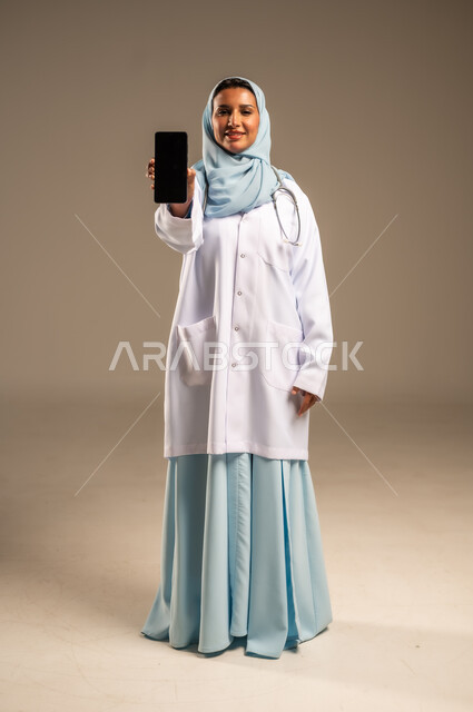 Providing services to patients efficiently, using technology in research and development, conducting medical consultations remotely, portrait of a Saudi Arabian Gulf female doctor wearing a colorful abaya with a white coat working on a mobile phone, health professions and jobs, colorful background