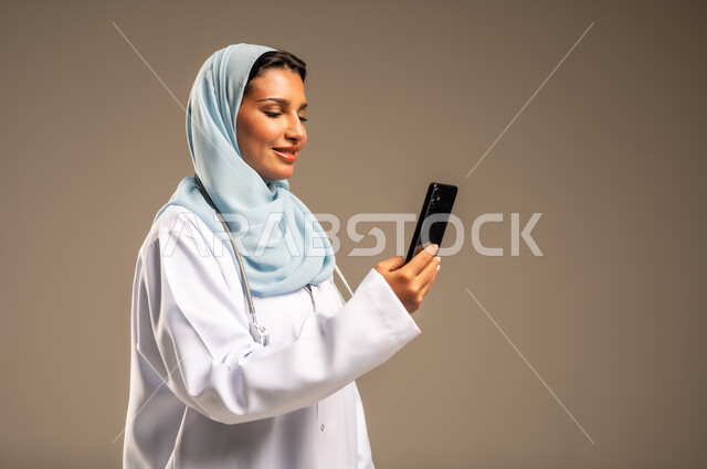 Providing services to patients efficiently, using technology in research and development, conducting medical consultations remotely, portrait of a Saudi Arabian Gulf female doctor wearing a colorful abaya with a white coat working on a mobile phone, health professions and jobs, colorful background