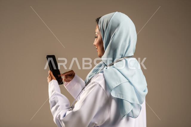 تقديم الخدمات للمرضى بكل كفاءه، استخدام التكنولوجيا في البحث والتطوير، اجراء الاستشارات الطبيه عن بعد، بورتريه لطبيبة عربية خليجية سعودية ترتدي عباءة ملونة مع المعطف الابيض تعمل على الهاتف المحمول، مهن ووظائف صحية، خلفيه ملونة