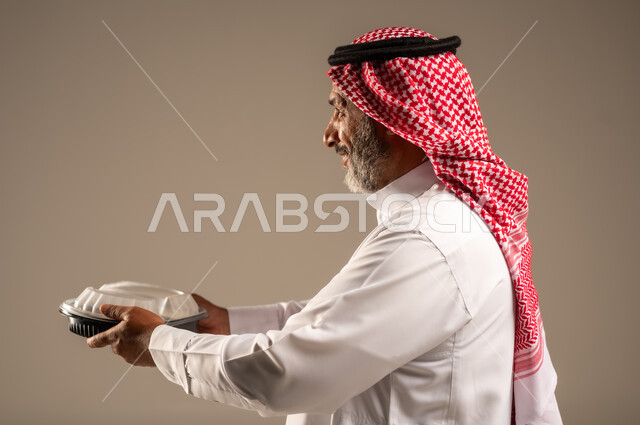 الاستمتاع بتناول الوجبات السريعة, بورتريه مقرب من الجانب لرجل كبير السن عربي خليجي سعودي مبتسم يرتدي الشماغ والثوب التقليدي يمسك بيده وجبة طعام، توفير الغذاء للمحتاجين، مبادرات وتبرعات انسانية خيرية، خلفية بيج