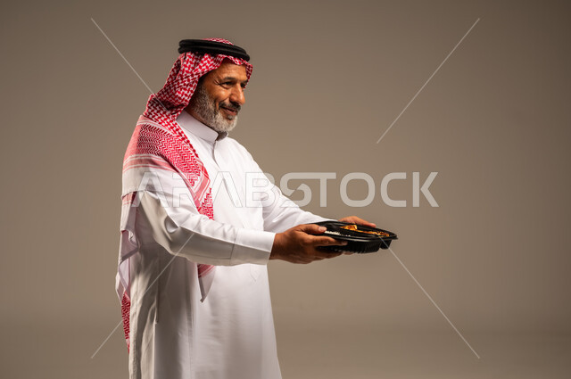الاستمتاع بتناول الوجبات السريعة, بورتريه مقرب من الجانب لرجل كبير السن عربي خليجي سعودي مبتسم يرتدي الشماغ والثوب التقليدي يمسك بيده وجبة طعام، توفير الغذاء للمحتاجين، مبادرات وتبرعات انسانية خيرية، خلفية بيج