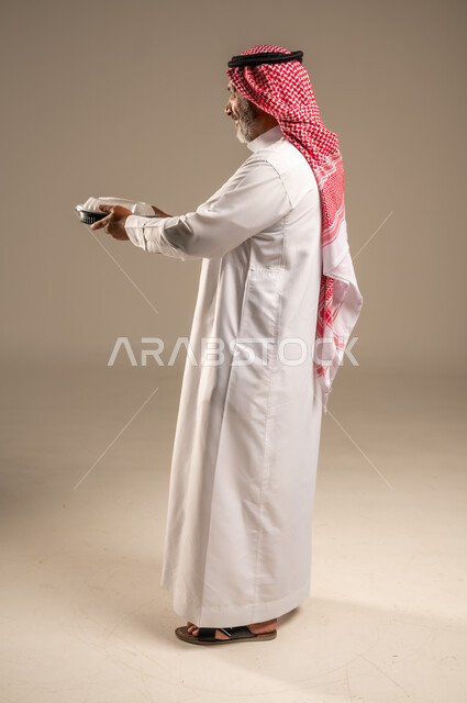 مبادرات وتبرعات انسانية خيرية، الاستمتاع بتناول الوجبات السريعة, بورتريه من الجانب لرجل كبير السن عربي خليجي سعودي مبتسم يرتدي الشماغ والثوب التقليدي يمسك بيده وجبة طعام، توفير الغذاء للمحتاجين، صورة الجسم بالطول الكامل، خلفية بيج