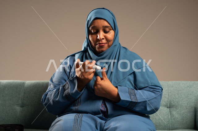 الفحص الطبي السريع في المنزل، أجهزة إلكترونية حديثة ومتطورة لمرضى داء السكري، بورتريه مقرب لامراة عربية خليجية سعودية مريضة ترتدي العباءة والحجاب تحمل جهاز قياس سكر الدم تقوم بفحص نسبة السكر في دمها، خلفية بيج