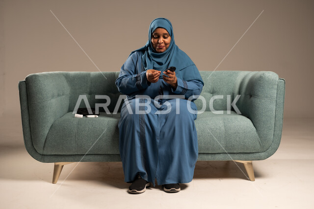 الفحص الطبي السريع في المنزل، أجهزة إلكترونية حديثة ومتطورة لمرضى داء السكري، بورتريه لامراة عربية خليجية سعودية مريضة ترتدي العباءة والحجاب تحمل جهاز قياس سكر الدم تقوم بفحص نسبة السكر في دمها، خلفية بيج