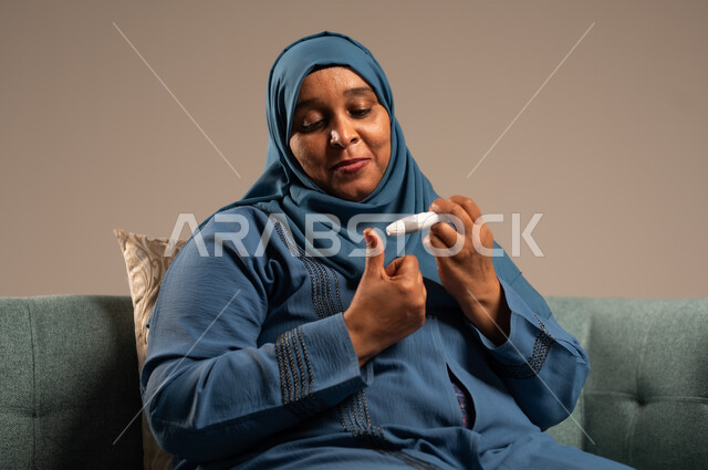 الفحص الطبي السريع في المنزل، أجهزة إلكترونية حديثة ومتطورة لمرضى داء السكري، بورتريه مقرب لامراة عربية خليجية سعودية مريضة ترتدي العباءة والحجاب تحمل جهاز قياس سكر الدم تقوم بفحص نسبة السكر في دمها، خلفية بيج