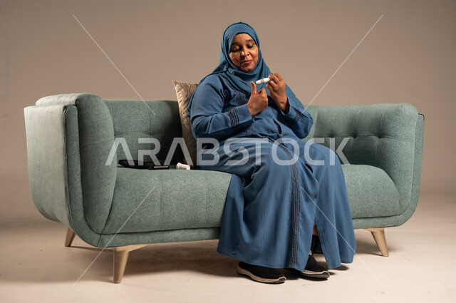 الفحص الطبي السريع في المنزل، أجهزة إلكترونية حديثة ومتطورة لمرضى داء السكري، بورتريه لامراة عربية خليجية سعودية مريضة ترتدي العباءة والحجاب تحمل جهاز قياس سكر الدم تقوم بفحص نسبة السكر في دمها، خلفية بيج