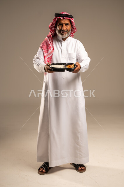 مبادرات وتبرعات انسانية خيرية، الاستمتاع بتناول الوجبات السريعة, بورتريه لرجل كبير السن عربي خليجي سعودي مبتسم يرتدي الشماغ والثوب التقليدي يمسك بيده وجبة طعام ينظر الى الكاميرا بإيماءات السرور، توفير الغذاء للمحتاجين، صورة الجسم بالطول الكامل، خلفية بيج