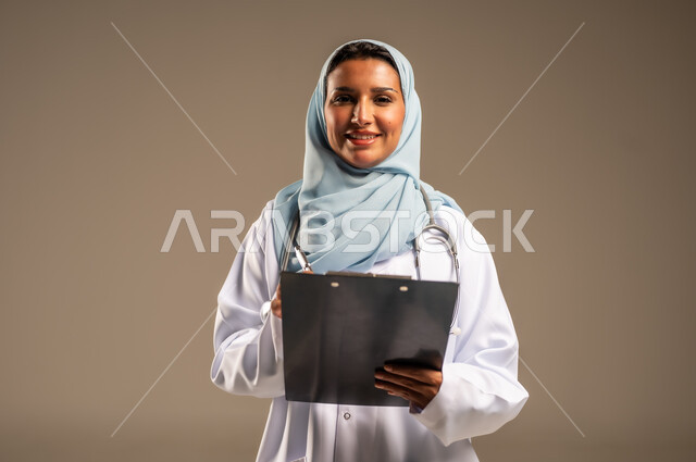 تقديم الخدمات والاستشارات الطبية للمراجعين، مفهوم الطب والرعاية الصحية، بورتريه لطبيبة عربية خليجية سعودية ترتدي عباءة ملونة مع المعطف الابيض تقوم بتدوين المعلومات العامة عن حالة المريض، مهن ووظائف نسائية، دراسة و مراجعة حالات المرضى، خلفيه ملونة