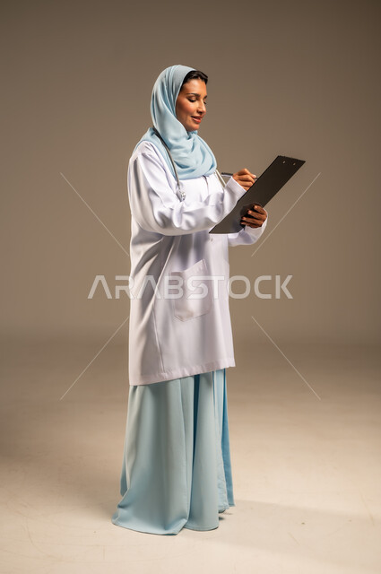 تقديم الخدمات والاستشارات الطبية للمراجعين، مفهوم الطب والرعاية الصحية، بورتريه لطبيبة عربية خليجية سعودية ترتدي عباءة ملونة مع المعطف الابيض تقوم بتدوين المعلومات العامة عن حالة المريض، مهن ووظائف نسائية، دراسة و مراجعة حالات المرضى، خلفيه ملونة