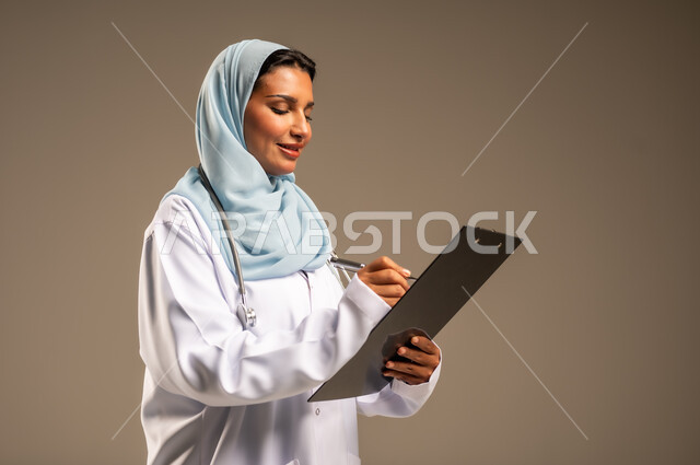 تقديم الخدمات والاستشارات الطبية للمراجعين، مفهوم الطب والرعاية الصحية، بورتريه لطبيبة عربية خليجية سعودية ترتدي عباءة ملونة مع المعطف الابيض تقوم بتدوين المعلومات العامة عن حالة المريض، مهن ووظائف نسائية، دراسة و مراجعة حالات المرضى، خلفيه ملونة