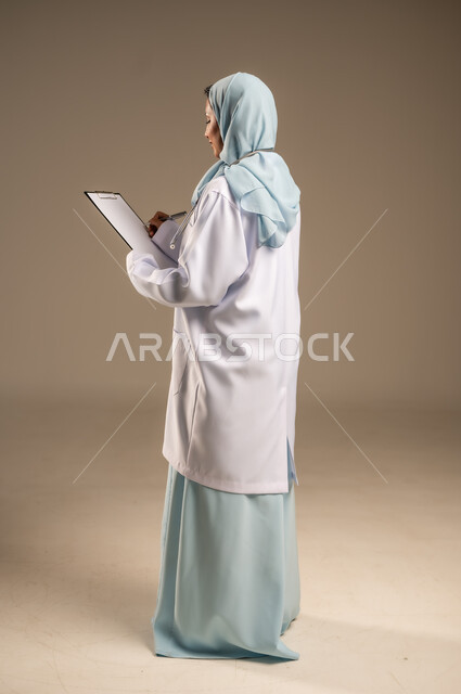 تقديم الخدمات والاستشارات الطبية للمراجعين، مفهوم الطب والرعاية الصحية، بورتريه لطبيبة عربية خليجية سعودية ترتدي عباءة ملونة مع المعطف الابيض تقوم بتدوين المعلومات العامة عن حالة المريض، مهن ووظائف نسائية، دراسة و مراجعة حالات المرضى، خلفيه ملونة