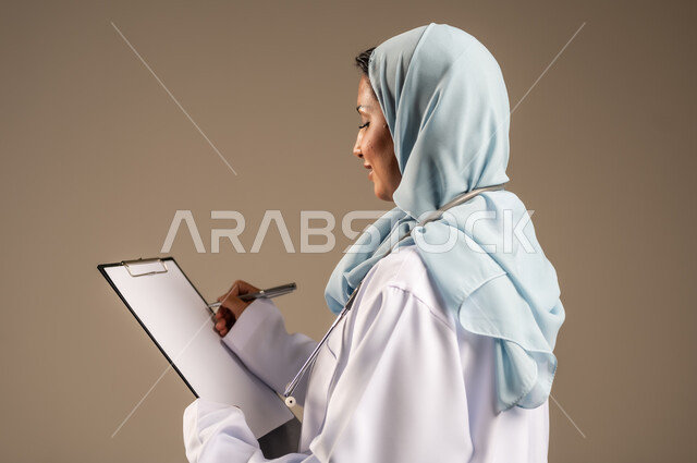 تقديم الخدمات والاستشارات الطبية للمراجعين، مفهوم الطب والرعاية الصحية، بورتريه لطبيبة عربية خليجية سعودية ترتدي عباءة ملونة مع المعطف الابيض تقوم بتدوين المعلومات العامة عن حالة المريض، مهن ووظائف نسائية، دراسة و مراجعة حالات المرضى، خلفيه ملونة