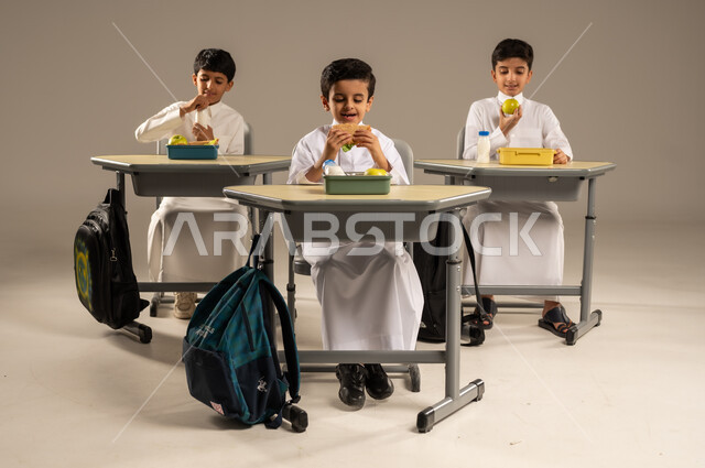 الوجبات الغنية بالعناصر الغذائية الهامة لبناء الجسم، الاستمتاع بتناول الأطعمة الصحية في وقت الفسحة المدرسية، موسم العودة للمدارس، بورتريه لمجموعة من الطلاب العربيين الخليجيين السعوديين يرتدون الزي المدرسي يجلسون في مقاعدهم وأمامهم اللانش البوكس، خلفية رمادية