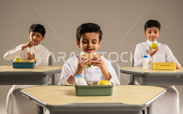 الوجبات الغنية بالعناصر الغذائية الهامة لبناء الجسم، الاستمتاع بتناول الأطعمة الصحية في وقت الفسحة المدرسية، موسم العودة للمدارس، بورتريه لمجموعة من الطلاب العربيين الخليجيين السعوديين يرتدون الزي المدرسي يجلسون في مقاعدهم وأمامهم اللانش البوكس، خلفية رمادية