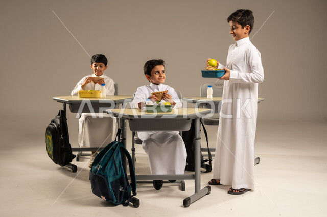 الوجبات الغنية بالعناصر الغذائية الهامة لبناء الجسم، الاستمتاع بتناول الأطعمة الصحية في وقت الفسحة المدرسية، موسم العودة للمدارس، بورتريه لمجموعة من الطلاب العربيين الخليجيين السعوديين يرتدون الزي المدرسي يجلسون في مقاعدهم وأمامهم اللانش البوكس، خلفية رمادية