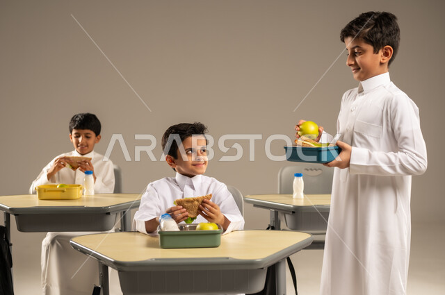 الوجبات الغنية بالعناصر الغذائية الهامة لبناء الجسم، الاستمتاع بتناول الأطعمة الصحية في وقت الفسحة المدرسية، موسم العودة للمدارس، بورتريه لمجموعة من الطلاب العربيين الخليجيين السعوديين يرتدون الزي المدرسي يجلسون في مقاعدهم وأمامهم اللانش البوكس، خلفية رمادية