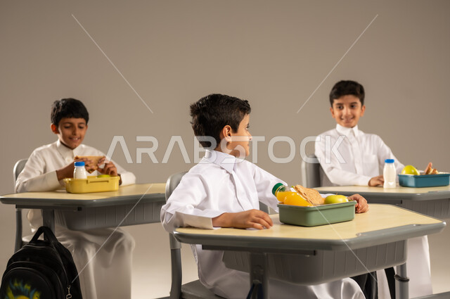 Meals rich in important nutrients for building the body, enjoying eating healthy foods during school break, back to school season, portrait of a group of Saudi Arabian Gulf students wearing school uniforms sitting in their seats with lunch boxes in front of them, gray background