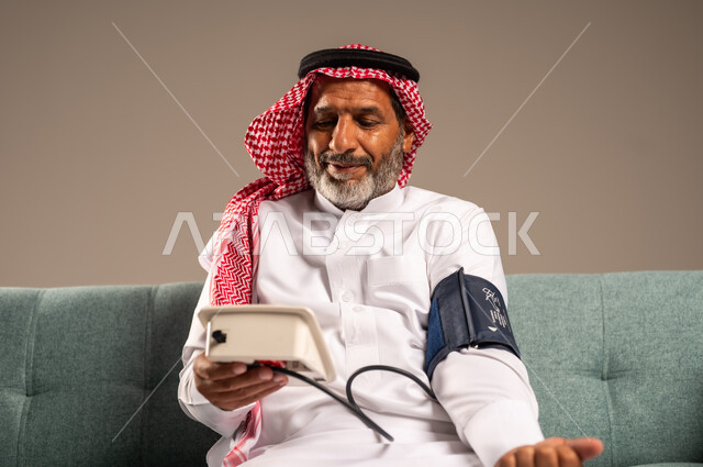 Looking at the device with gestures of pleasure, using the modern and advanced electronic scale for modern quick home examination, portrait of an elderly Saudi Gulf man wearing traditional thobe and shemagh sitting on a comfortable sofa measuring blood pressure alone, concept of medicine and health care, beige background
