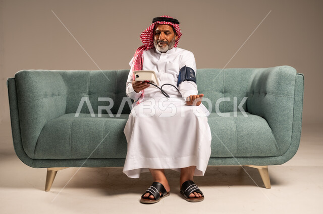 Looking at the device with gestures of pleasure, using the modern and advanced electronic scale for modern quick home examination, portrait of an elderly Saudi Gulf man wearing traditional thobe and shemagh sitting on a comfortable sofa measuring blood pressure alone, concept of medicine and health care, beige background