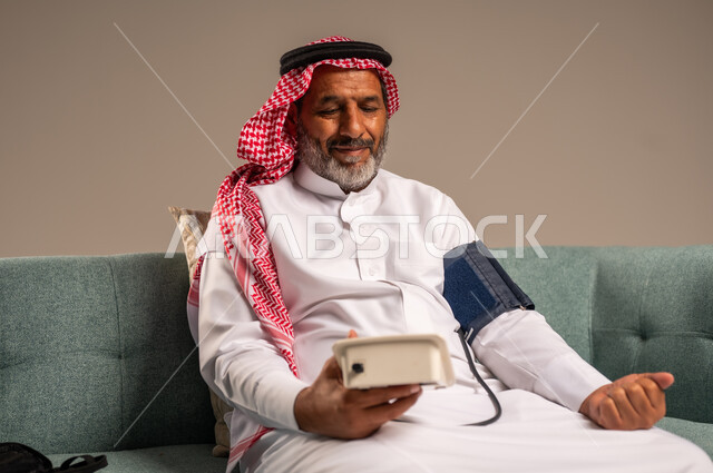 استخدام الميزان الاكتروني الحديث والمتطور للفحص المنزلي السريع الحديث، بورتريه لرجل مسن سعودي خليجي يرتدي الثوب التقليدي والشماغ يجلس على كنبة مريحة يقوم بقياس ضغط الدم لوحده، مفهوم الطب والرعاية الصحية، النظر الى الكاميرا بايماءات السرور، خلفية بيج