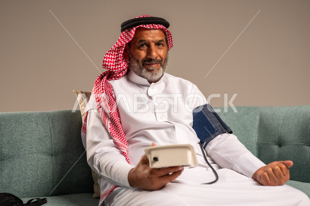 استخدام الميزان الاكتروني الحديث والمتطور للفحص المنزلي السريع الحديث، بورتريه لرجل مسن سعودي خليجي يرتدي الثوب التقليدي والشماغ يجلس على كنبة مريحة يقوم بقياس ضغط الدم لوحده، مفهوم الطب والرعاية الصحية، النظر الى الكاميرا بايماءات السرور، خلفية بيج