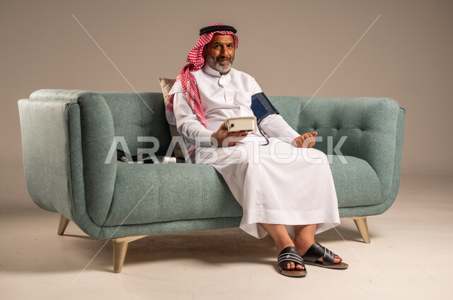 Using modern and advanced electronic scale for modern quick home examination, portrait of an elderly Saudi Gulf man wearing traditional thobe and shemagh sitting on a comfortable sofa measuring blood pressure by himself, concept of medicine and health care, looking at the camera with gestures of pleasure, beige background