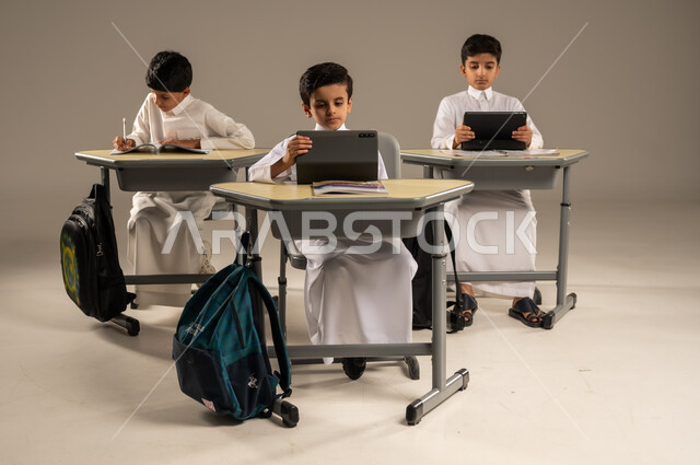 الاستفادة من التقنيات والتكنولوجيا الحديثة في التعلم والدراسة، المناهج المطورة والتعليم الالكتروني، تطور طرائق التدريس في مدارس المملكة، بورتريه لطلاب عربيين خليجيين سعوديين يرتدون الزي المدرسي يجلسون في مقاعدهم وأمامهم أجهزة التابلت المحمولة، خلفية رمادية
