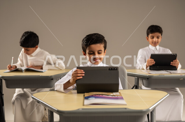 الاستفادة من التقنيات والتكنولوجيا الحديثة في التعلم والدراسة، المناهج المطورة والتعليم الالكتروني، تطور طرائق التدريس في مدارس المملكة، بورتريه لطلاب عربيين خليجيين سعوديين يرتدون الزي المدرسي يجلسون في مقاعدهم وأمامهم أجهزة التابلت المحمولة، خلفية رمادية