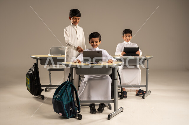 Benefiting from modern techniques and technology in learning and studying, developed curricula and e-learning, development of teaching methods in the Kingdom's schools, portrait of Saudi Arabian Gulf students wearing school uniforms sitting in their seats with portable tablets in front of them, gray background