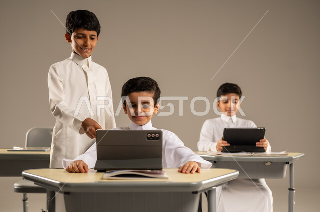 Benefiting from modern techniques and technology in learning and studying, developed curricula and e-learning, development of teaching methods in the Kingdom's schools, portrait of Saudi Arabian Gulf students wearing school uniforms sitting in their seats with portable tablets in front of them, gray background