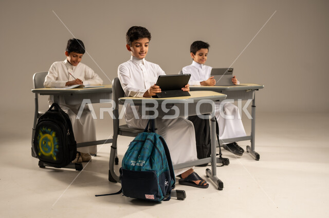 Developed curricula and e-learning, benefiting from modern techniques and technology in learning and studying, development of teaching methods in the Kingdom's schools, portrait of Saudi Arabian Gulf students wearing school uniforms sitting in their seats with portable tablets in front of them, gray background