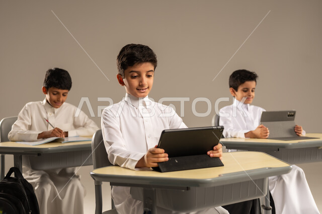 المناهج المطورة والتعليم الالكتروني، الاستفادة من التقنيات والتكنولوجيا الحديثة في التعلم والدراسة، تطور طرائق التدريس في مدارس المملكة، بورتريه لطلاب عربيين خليجيين سعوديين يرتدون الزي المدرسي يجلسون في مقاعدهم وأمامهم أجهزة التابلت المحمولة، خلفية رمادية