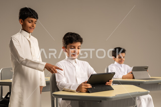 المناهج المطورة والتعليم الالكتروني، الاستفادة من التقنيات والتكنولوجيا الحديثة في التعلم والدراسة، تطور طرائق التدريس في مدارس المملكة، بورتريه لطلاب عربيين خليجيين سعوديين يرتدون الزي المدرسي يجلسون في مقاعدهم وأمامهم أجهزة التابلت المحمولة، خلفية رمادية