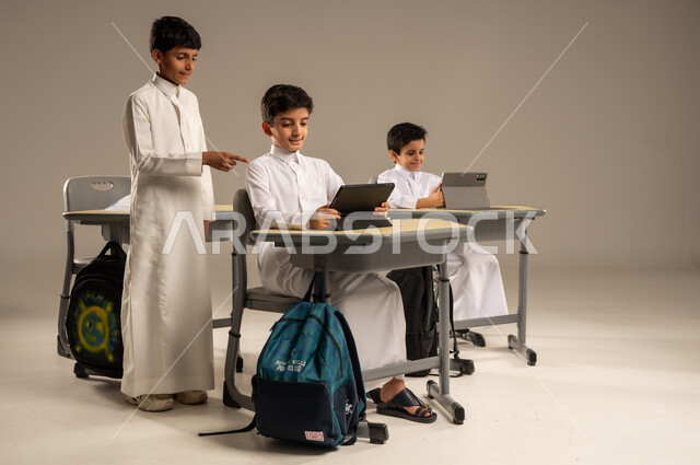المناهج المطورة والتعليم الالكتروني، الاستفادة من التقنيات والتكنولوجيا الحديثة في التعلم والدراسة، تطور طرائق التدريس في مدارس المملكة، بورتريه لطلاب عربيين خليجيين سعوديين يرتدون الزي المدرسي يجلسون في مقاعدهم وأمامهم أجهزة التابلت المحمولة، خلفية رمادية