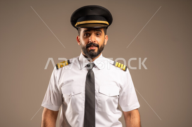  طاقم الاستقبال والترحيب بالركاب، العمل في مجال الطيران، مفهوم السياحة والسفر، رحلات جوية سياحية, خطوط الطيران السعودية,  بورتريه لكابتن طيار عربي سعودي خليجي مبتسم يرتدي الزي الرسمي يقف باستقامة ينظر الى الكاميرا بتعابير السعادة والسرور، خلفية ملونة