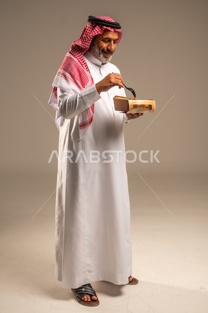 وصفات المأكولات الشهية، الاستمتاع بتناول الوجبات السريعة, بورتريه من الجانب لرجل كبير السن عربي خليجي سعودي مبتسم يرتدي الشماغ والثوب التقليدي يمسك بيده وجبة طعام، خدمات طلب واستلام الديليفري, صورة الجسم بالطول الكامل, خلفية بيج