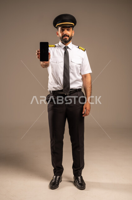 استخدام الاجهزة التقنية الحديثة، طاقم الاستقبال والترحيب بالركاب، العمل في مجال الطيران، مفهوم السياحة والسفر، رحلات جوية سياحية, خطوط الطيران السعودية,  بورتريه لكابتن طيار عربي سعودي خليجي مبتسم يرتدي الزي الرسمي يمسك بيده الهاتف المحمول، خلفية ملونة