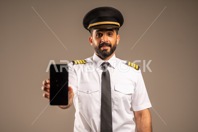 استخدام الاجهزة التقنية الحديثة، طاقم الاستقبال والترحيب بالركاب، العمل في مجال الطيران، مفهوم السياحة والسفر، رحلات جوية سياحية, خطوط الطيران السعودية,  بورتريه لكابتن طيار عربي سعودي خليجي مبتسم يرتدي الزي الرسمي يمسك بيده الهاتف المحمول، خلفية ملونة