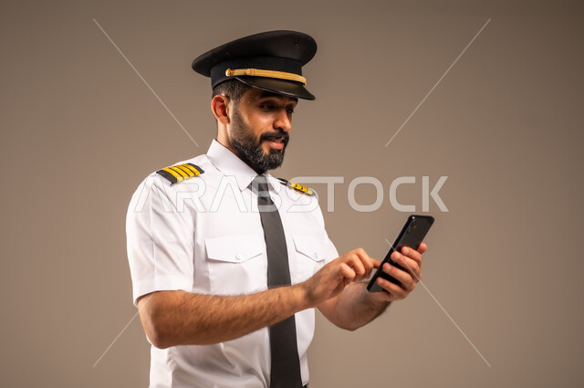 استخدام الاجهزة التقنية الحديثة، طاقم الاستقبال والترحيب بالركاب، العمل في مجال الطيران، مفهوم السياحة والسفر، رحلات جوية سياحية, خطوط الطيران السعودية,  بورتريه لكابتن طيار عربي سعودي خليجي مبتسم يرتدي الزي الرسمي يمسك بيده الهاتف المحمول، خلفية ملونة